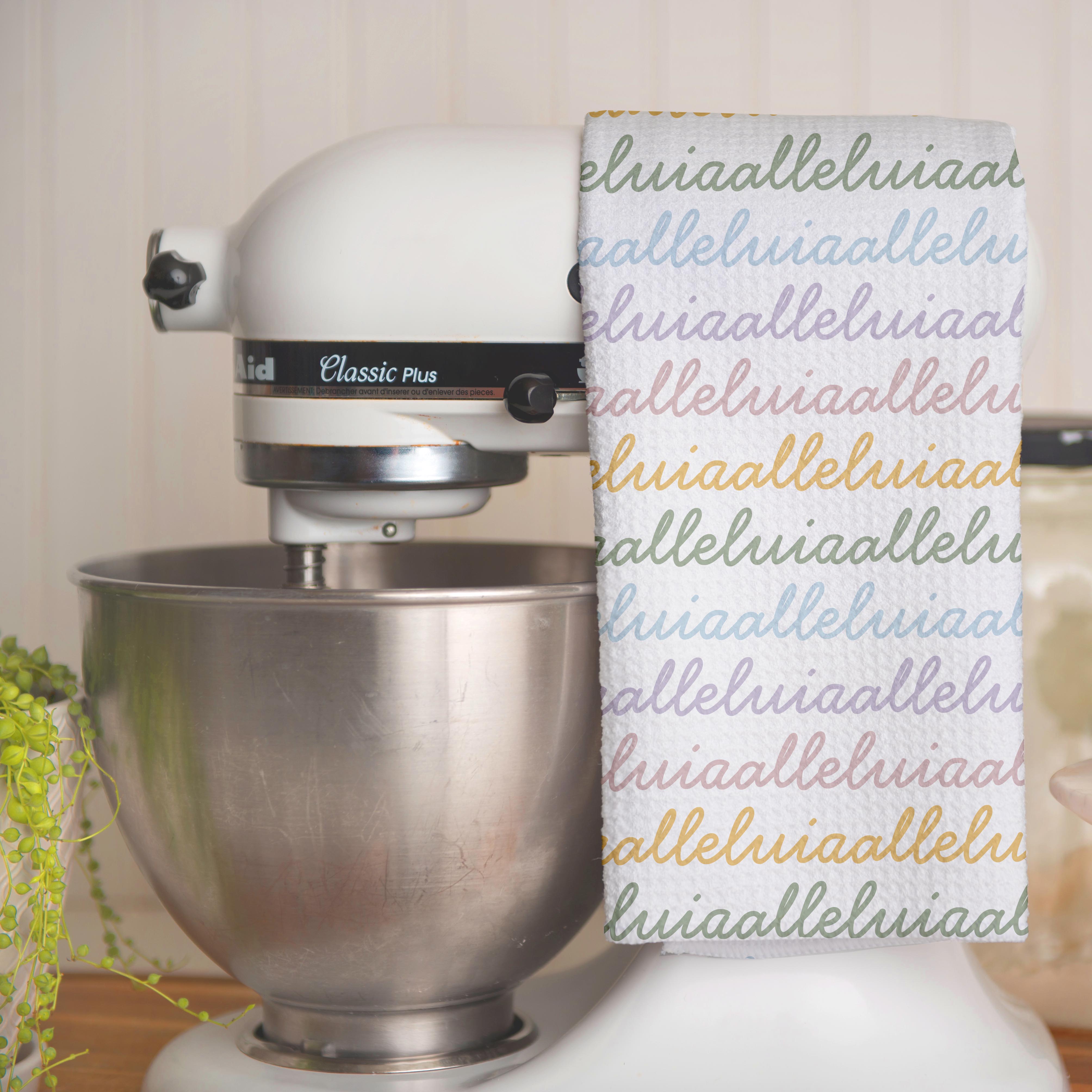 White colored, waffle textured dish towel folded in half, hanging on a white kitchen mixer, with the design that says alleluia, alleluia repeatedly in cursive writing, in colors pastel pink, pastel yellow, pastel blue and pastel green.