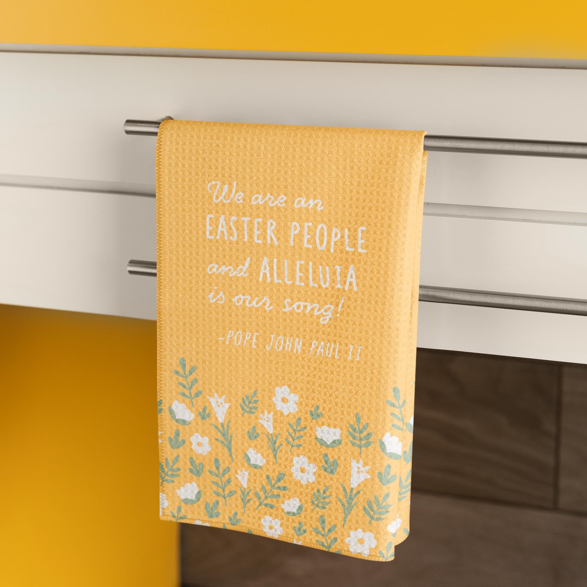 Yellow, waffle textured dish towel, folded in half, showing the design of subtle white flowers and pale green leaves with the wordings that say "We are an EASTER PEOPLE and ALLELUIA is our song! -Pope John Paul II"